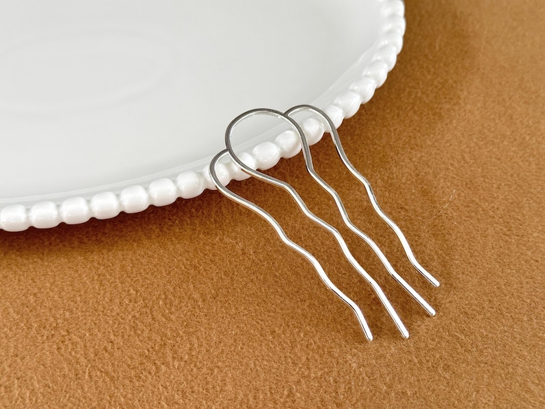 May include: A silver-colored hair comb with three wavy prongs, resting on a white plate with a beaded edge. The background is a solid brown color.