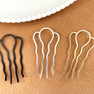 May include: Three hair pins in black, silver, and gold. Each pin has a wavy design with a rounded top and three prongs. The pins are displayed on a brown surface with a white plate in the background.