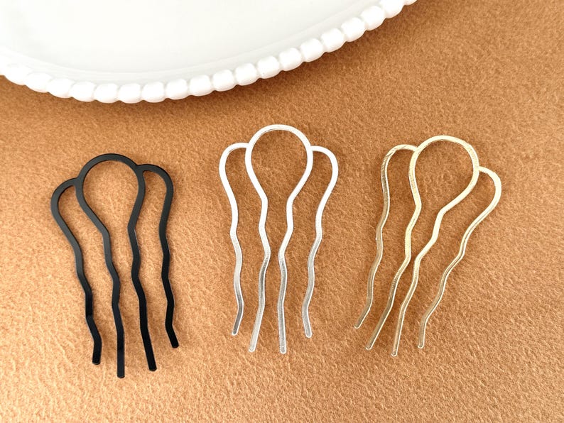 May include: Three hair pins in black, silver, and gold. Each pin has a wavy design with a rounded top and three prongs. The pins are displayed on a brown surface with a white plate in the background.