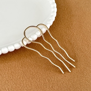 May include: Gold-toned hair combs with a wavy design. The hair accessories are arranged on a brown surface next to a white plate with a beaded edge. These combs are ideal for adding a touch of elegance to any hairstyle.
