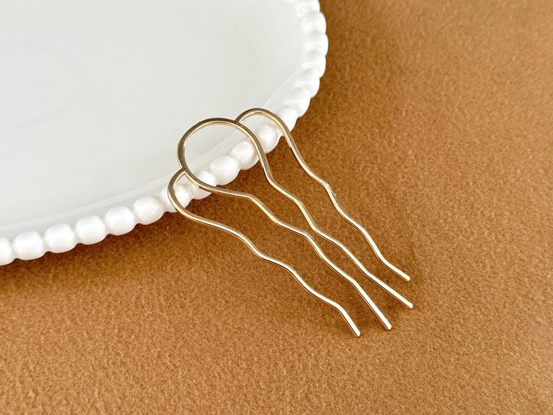 May include: Gold-toned hair combs with a wavy design. The hair accessories are arranged on a brown surface next to a white plate with a beaded edge. These combs are ideal for adding a touch of elegance to any hairstyle.