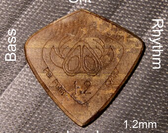 Könnte beinhalten: Ein braunes, dreieckiges Gitarrenplektrum mit einer Dicke von 1,2 mm. Das Plektrum hat ein Design eines stilisierten Krebses mit dem Text "Pal Fender" und "1.2" darauf. Das Plektrum ist mit den Wörtern "Grit", "Rhythm", "Bass", "Lead" beschriftet.