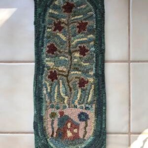 May include: A hand-woven rug with a green border and an oval-shaped center. The center features a stylized tree with red leaves and a small house at the base. The rug is made with a variety of colors, including green, blue, red, and yellow.