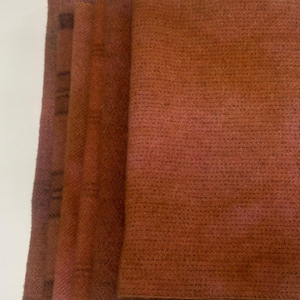 May include: A stack of four folded, textured, rust-colored fabric squares. The fabric has a woven appearance with a subtle gradient of color, transitioning from darker shades to lighter hues. The edges of the fabric are visible.