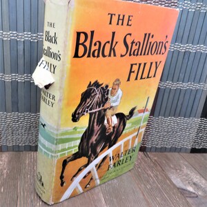 Puede incluir: Una cubierta de libro vintage con fondo naranja que presenta el t&iacute;tulo "The Black Stallion's Filly" en texto negro. La cubierta representa un caballo negro corriendo en una pista con un jinete mont&aacute;ndolo.