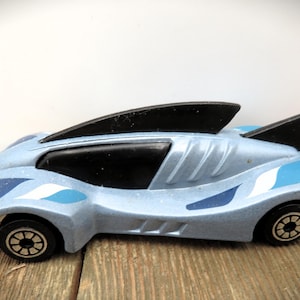 May include: A light blue toy car with black fins and black wheels. The car has a futuristic design with blue and white accents. The toy car is sitting on a wooden surface.
