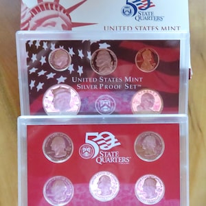 May include: A United States Mint Silver Proof Set in a clear plastic case. The set includes various coins, including quarters and pennies, displayed against a red and white background. The packaging features the Statue of Liberty and the text "50 State Quarters".