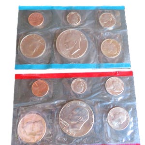 May include: Two sealed coin sets, each containing a selection of U.S. coins, including pennies, nickels, dimes, quarters, and half dollars. The sets are in clear plastic with blue and red sealed edges. The half dollars are dated 1971.