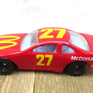 May include: A red toy race car featuring yellow number 27 decals on the roof and side doors. The bonnet displays a yellow McDonald's logo, while the rear quarter panel features the McDonald's text and logo. The car has black wheels and windows.