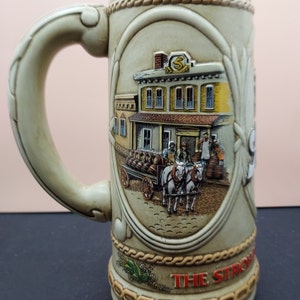 May include: A ceramic beer stein with a beige background and a detailed illustration of a building with a sign that reads "The Stroh's". The building is surrounded by a scene of a horse-drawn wagon carrying barrels.