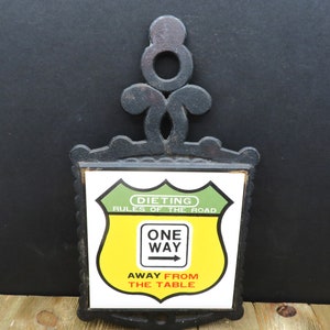 May include: Black cast iron hot pad with a white ceramic tile featuring a yellow and green road sign that reads "Dieting Rules of the Road" with a smaller sign that reads "One Way Away From The Table".