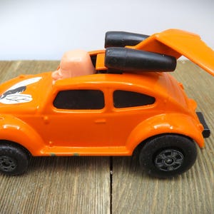 May include: Orange toy car with black wheels and a rear wing. The car has a light-colored figure in the driver's seat and a white and black design on the hood. The toy car is on a wooden surface.