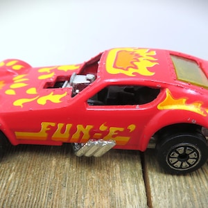 May include: A red toy car with yellow flame designs on the hood and roof. The car has the word "FUN-E" in yellow and orange lettering on the side. Black wheels and a silver engine are visible.