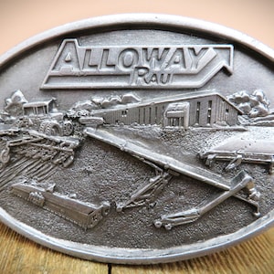 May include: A silver belt buckle with a 3D design of a farm scene. The buckle features a tractor, a building with the text "Alloway Rau", and a conveyor belt.