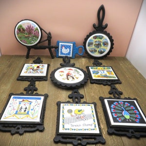 May include: Nine vintage cast iron hot pads with colorful ceramic tiles. The tiles feature various designs including a rooster, a peacock, a tennis player, a couple dancing, a spinning wheel, and a scene from Yellowstone Park.