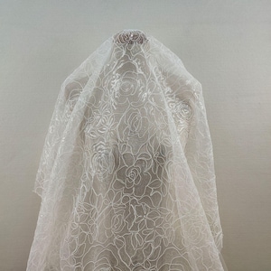 Può includere: Tessuto bianco trasparente con un motivo floreale ricamato. Il tessuto è drappeggiato su un manichino.