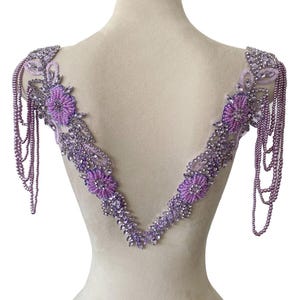 May include: A V-shaped lavender beaded applique featuring floral motifs, clear crystals, and dangling pearl-like bead strands on the shoulders. The design is composed of intricate lavender seed beads, silver-toned accents, and sparkling crystal embellishments.
