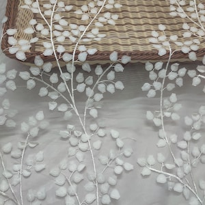 Peut inclure: Tissu en dentelle florale blanche avec un motif de feuilles délicat. Les feuilles sont en relief et texturées, créant un effet tridimensionnel. Le tissu est posé sur un fond neutre, avec un panier en osier en arrière-plan.