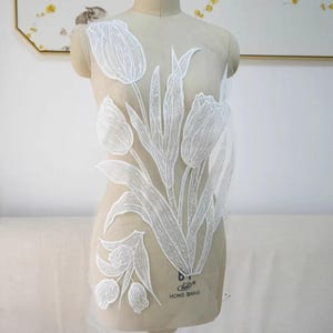 Puede incluir: Aplique de encaje blanco con cuentas que presenta un diseño de tulipes con tallos y hojas. El aplique se muestra en un maniquí blanco.
