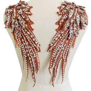May include: Two ornate, copper-colored shoulder pieces, resembling wings, are adorned with shimmering crystals and faux pearls. The intricate design features a layered, textured appearance, perfect for embellishing garments or costumes.