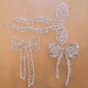 May include: Three white and silver beaded embellishments in the shape of bows. The bows have long tails and are perfect for adding a touch of sparkle to clothing or crafts.