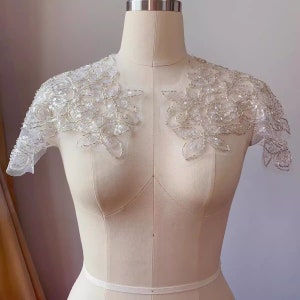 Può includere: Una stola da sposa in pizzo bianco perlato con maniche corte. La stola è realizzata in pizzo delicato con un motivo floreale ed è decorata con perline scintillanti.