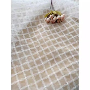 May include: White tulle fabric with a grid pattern of small, gathered circles. The fabric is sheer and delicate, with a soft, flowing texture.