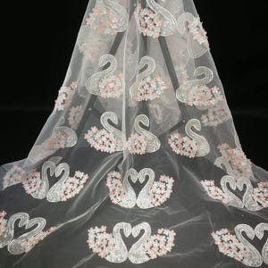 May include: White tulle fabric with embroidered white swans and pink flowers. The fabric is sheer and delicate, with a soft, romantic feel.