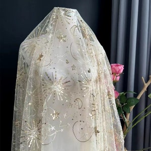 Può includere: Un tessuto di tulle avorio trasparente con paillettes dorate in un motivo celeste di stelle, lune e raggi di sole.