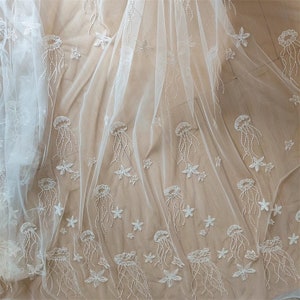 Pode incluir: Tecido branco transparente com desenhos bordados de medusas e estrelas do mar. O tecido é delicado e tem uma textura macia e fluida.