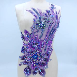 Puede incluir: Aplique morado y plateado con diseño floral. El aplique presenta cristales grandes y brillantes y cuentas iridiscentes más pequeñas dispuestas en un patrón simétrico. El diseño está sobre un fondo transparente de color lavanda.