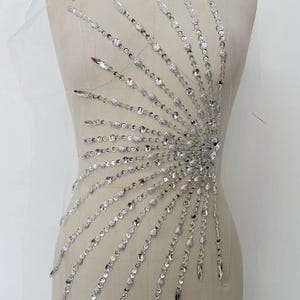 May include: A silver rhinestone applique with a sunburst design. The applique is made of small, round rhinestones and larger, teardrop-shaped rhinestones.