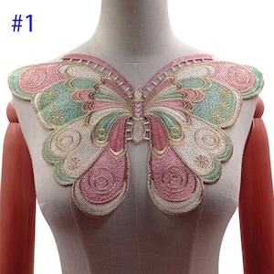 Può includere: Un'applicazione a forma di farfalla ricamata in rosa, verde e oro. La farfalla ha un design dettagliato con volute e cerchi. L'applicazione è perfetta per aggiungere un tocco di fantasia agli abiti o ai lavori artigianali.