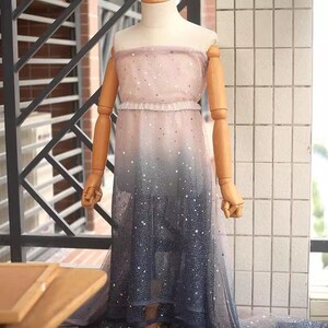 K&ouml;nnte beinhalten: Ein tr&auml;gerloses Ombre-Kleid auf einer Schaufensterpuppe, das von Rosa zu Marineblau &uuml;bergeht. Das Kleid ist mit silbernen Sternen und einem R&uuml;schendetail an der Taille verziert. Der Stoff wirkt transparent und flie&szlig;end. Geeignet f&uuml;r besondere Anl&auml;sse.