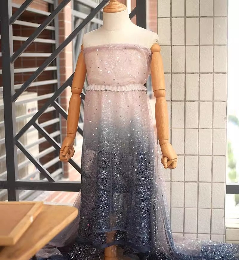 K&ouml;nnte beinhalten: Ein tr&auml;gerloses Ombre-Kleid auf einer Schaufensterpuppe, das von Rosa zu Marineblau &uuml;bergeht. Das Kleid ist mit silbernen Sternen und einem R&uuml;schendetail an der Taille verziert. Der Stoff wirkt transparent und flie&szlig;end. Geeignet f&uuml;r besondere Anl&auml;sse.
