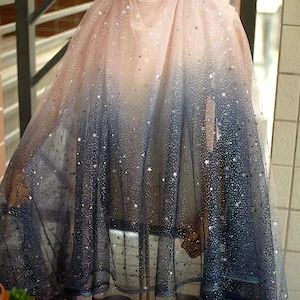 Könnte beinhalten: Ein durchsichtiger Stoff mit Farbverlauf, der von zartem Rosa zu tiefem Marineblau übergeht. Der Stoff ist mit silbernen sternförmigen Pailletten verziert, die ein himmlisches Aussehen erzeugen. Das Material scheint Tüll zu sein, geeignet für Kleider oder Dekorationszwecke.