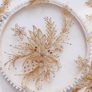 Puede incluir: Un delicado diseño de bordado floral en oro y blanco sobre un fondo blanco. El bordado presenta detalles intrincados y una variedad de texturas.