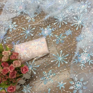 Op de afbeelding: Witte, doorschijnende stof met iriserende pailletten en blauwe sneeuwvlokken borduurwerk. De stof ligt op een houten oppervlak.
