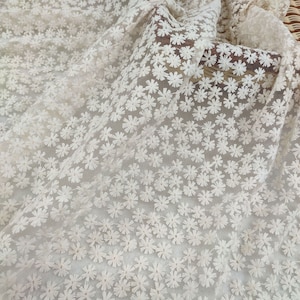 Può includere: Tessuto in pizzo floreale bianco sporco. Il materiale delicato è ricoperto da piccoli fiori ricamati a forma di margherita. Il tessuto è drappeggiato, mostrando la consistenza e la trasparenza del pizzo. Ideale per abiti da sposa, abiti o progetti artigianali.