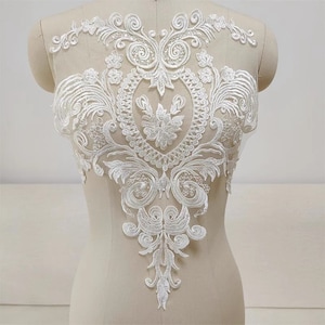 May include: White lace applique with a floral design. The lace is delicate and intricate, with a scalloped edge. This applique would be perfect for adding a touch of elegance to a wedding dress, bridal gown, or other special occasion garment.