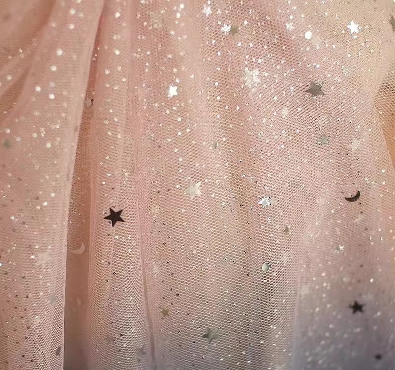 K&ouml;nnte beinhalten: Nahaufnahme eines hellrosa T&uuml;llstoffs mit zarter Netzstruktur. Der Stoff ist mit silbernen Glitzerpartikeln und sternf&ouml;rmigen Pailletten verziert, wodurch ein schimmernder Effekt entsteht. Die Gesamt&auml;sthetik ist weich und vertr&auml;umt.