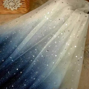 K&ouml;nnte beinhalten: Ein durchscheinender Stoff mit Farbverlauf, der von tiefem Marineblau zu Wei&szlig; &uuml;bergeht. Der Stoff ist mit silbernen Glitzersternen und Monden verziert und erzeugt ein himmlisches Thema. Ideal f&uuml;r Bastelarbeiten oder dekorative Zwecke.