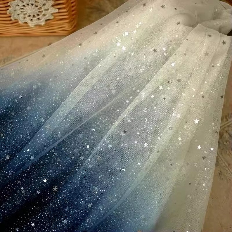 K&ouml;nnte beinhalten: Ein durchscheinender Stoff mit Farbverlauf, der von tiefem Marineblau zu Wei&szlig; &uuml;bergeht. Der Stoff ist mit silbernen Glitzersternen und Monden verziert und erzeugt ein himmlisches Thema. Ideal f&uuml;r Bastelarbeiten oder dekorative Zwecke.