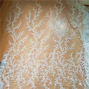 May include: White embroidered lace fabric with a delicate floral pattern. The lace is on a sheer mesh background.