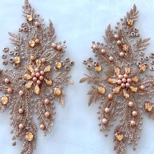 Könnte beinhalten: Zwei goldfarbene Spitzenapplikationen mit Perlen- und Strassbesatz. Jede Applikation zeigt ein florales Design mit einem zentralen Cluster aus Perlen und Strass.