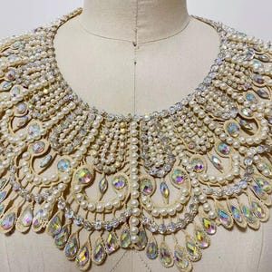 Puede incluir: Un collar grande, adornado, de color crema con un borde festoneado. El collar está hecho de perlas, pedrería y cristales transparentes.