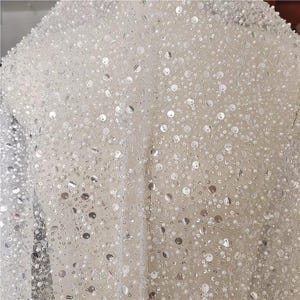 May include: A sheer white fabric overlay densely embellished with a combination of small round white beads and flat circular silver sequins. The intricate beadwork and sequins are scattered across the fine mesh netting, creating a reflective texture.