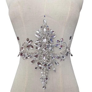 Puede incluir: Una decoración de pedrería plateada con forma de lágrima y una punta puntiaguda. La decoración está hecha de pequeñas piedras de pedrería transparentes y es perfecta para añadir brillo a la ropa o los accesorios.