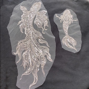 Könnte beinhalten: Zwei weiße Perlenapplikationen, eine mit einer Meerjungfrau und eine mit einem Fisch, auf einem weißen Netzstoff.