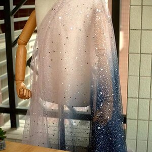 K&ouml;nnte beinhalten: Ein transparenter Stoff mit Farbverlauf, der von einem zarten Rosa zu einem tiefen Marineblau &uuml;bergeht. Der Stoff ist mit silbernen Sternen und Mondformen verziert, wodurch ein himmlisches Muster entsteht. Der Stoff ist &uuml;ber einer Schaufensterpuppe drapiert.